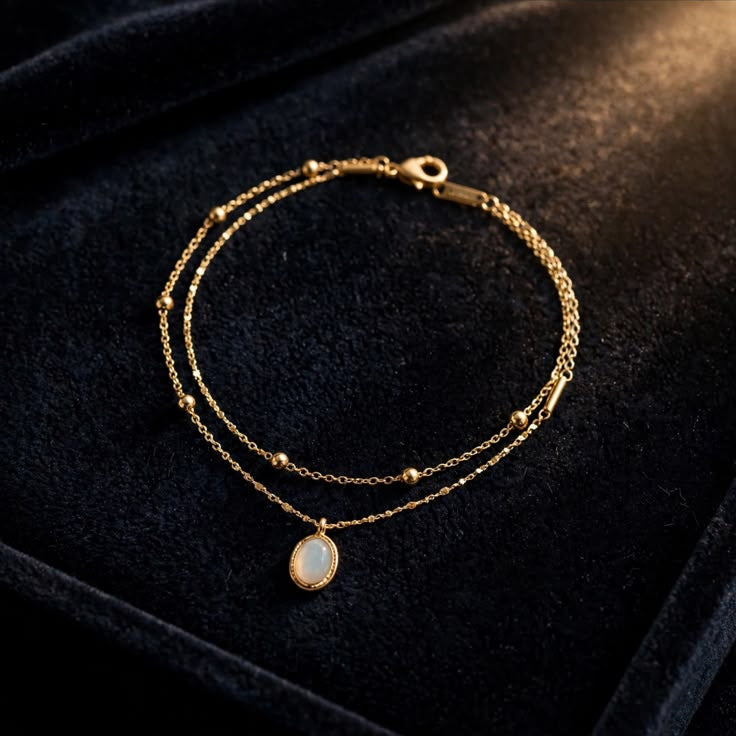 A delicate gold double-layered chain bracelet with a small teardrop-shaped white pearl pendant, displayed on a black velvet jewelry box.