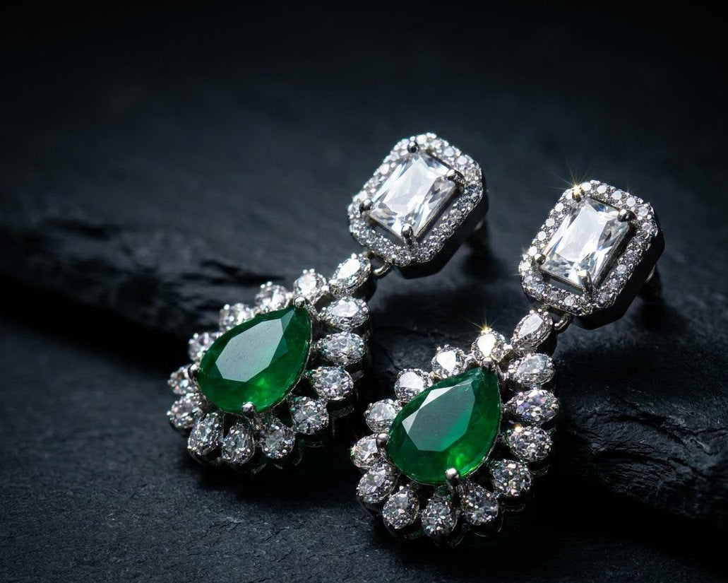 A pair of drop earrings featuring large emerald-cut green gemstones surrounded by clear stones in silver-toned settings, displayed on a dark textured surface.