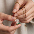 A close-up of a person's hands holding a delicate gold chain necklace with a flower-shaped pendant. The pendant features a white crystal center and green gemstone accents on the stem, set against a soft beige fabric background.