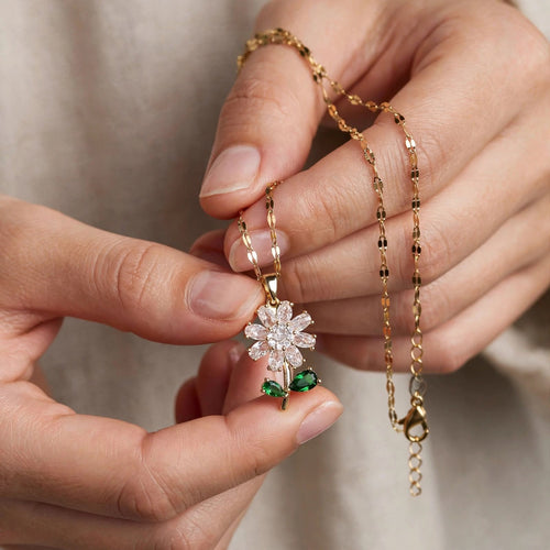 A close-up of a person's hands holding a delicate gold chain necklace with a flower-shaped pendant. The pendant features a white crystal center and green gemstone accents on the stem, set against a soft beige fabric background.