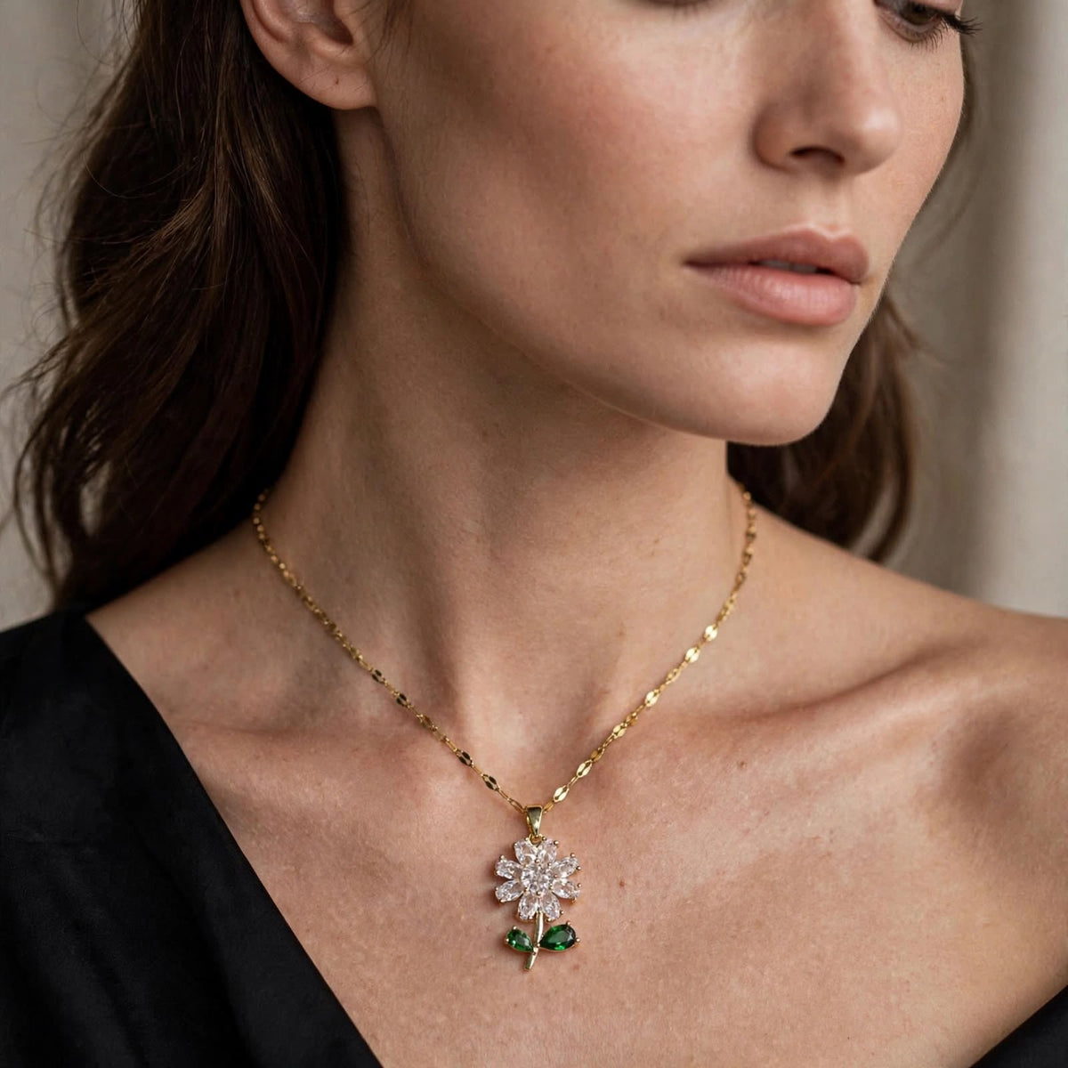 A close-up lifestyle shot of a model wearing a delicate gold chain necklace with a flower pendant. The pendant features a clear pavé diamond center with green emerald-cut gemstone leaves.