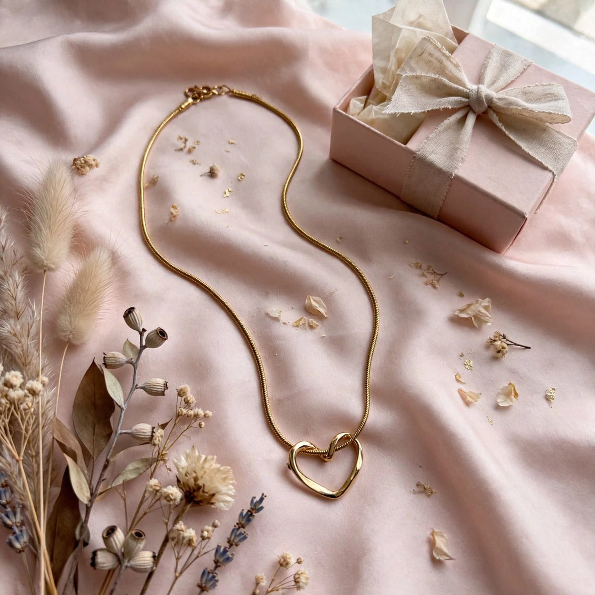 A gold-toned snake chain necklace with a double interlocking heart pendant, styled on a soft pink satin fabric alongside dried flowers and a pink gift box with a cream ribbon.