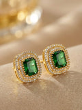 A pair of gold-toned stud earrings with rectangular green gemstones surrounded by double rows of small clear stones, displayed on a beige fabric surface with a softly blurred gold-toned background.