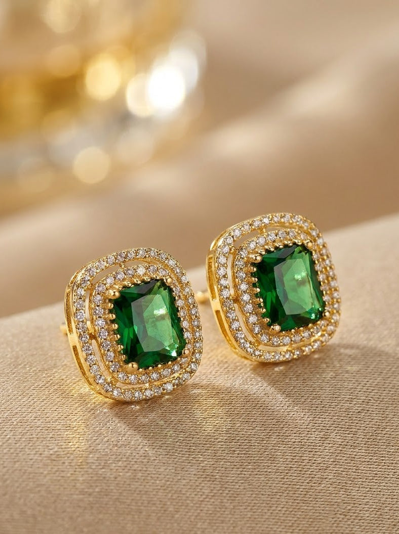 A pair of gold-toned stud earrings with rectangular green gemstones surrounded by double rows of small clear stones, displayed on a beige fabric surface with a softly blurred gold-toned background.