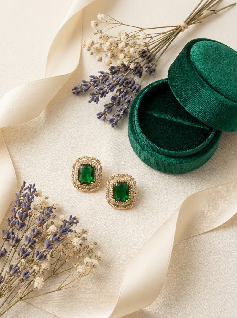 A pair of gold-toned square stud earrings with emerald-cut green stones, styled on a cream background with dried lavender, a cream ribbon, and an open dark green velvet box.