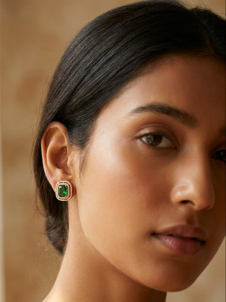 A close-up of a woman wearing a small gold stud earring with a rectangular green gemstone, set against a warm beige background.