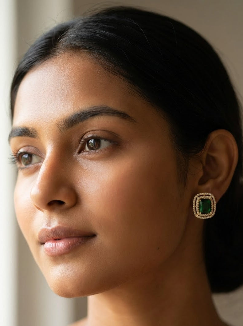 A close-up portrait of a woman wearing a single square green gemstone earring with a gold-toned border, shown against a soft neutral indoor background.