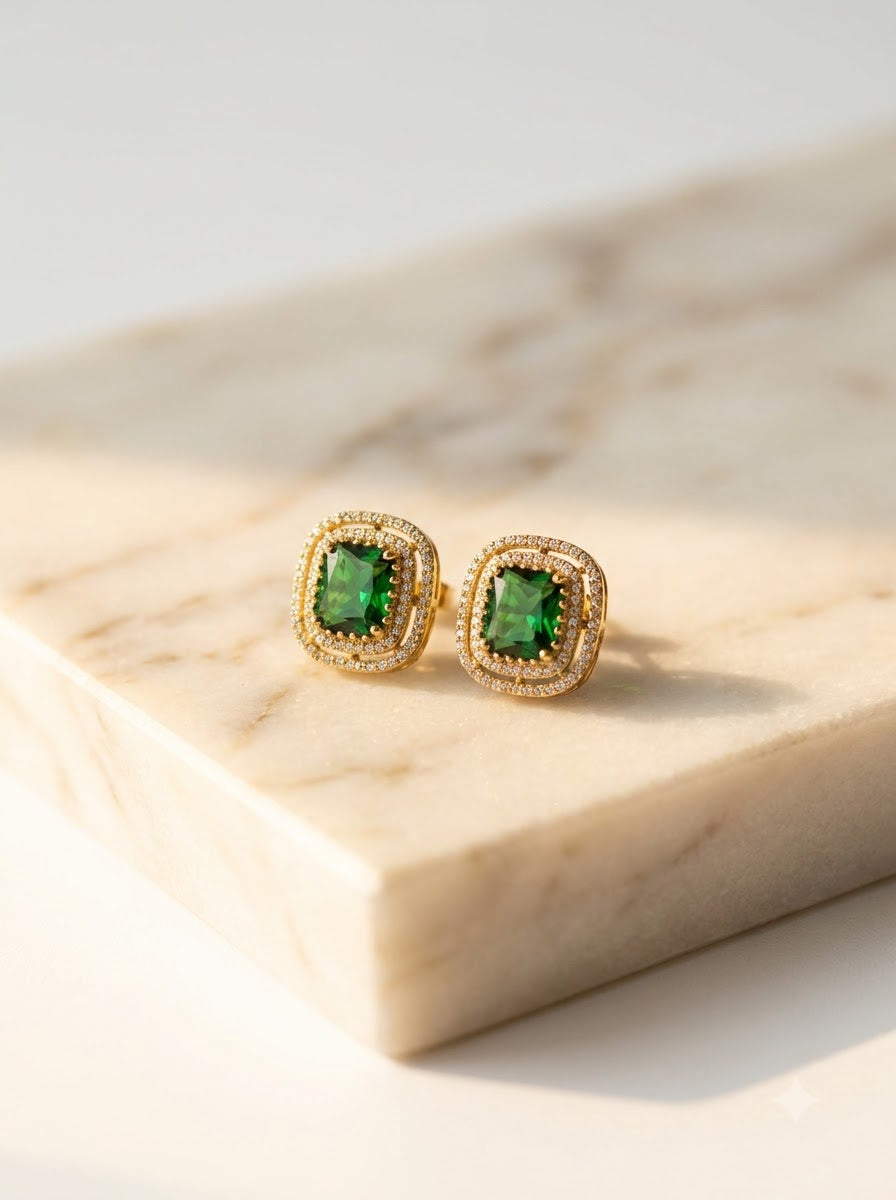 A pair of gold square stud earrings with green cushion-cut center stones and a halo of small clear stones, displayed on a light marble block.