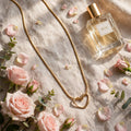 A gold heart-shaped pendant necklace and a clear glass perfume bottle labeled ROSE are arranged on a beige linen surface with pink roses, petals, and greenery in soft natural light.