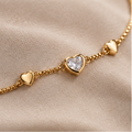 A collage showing a gold heart charm bracelet worn on a wrist and displayed flat. The bracelet features a delicate gold chain with multiple small gold hearts, including one larger heart with a clear heart-shaped crystal centerpiece.