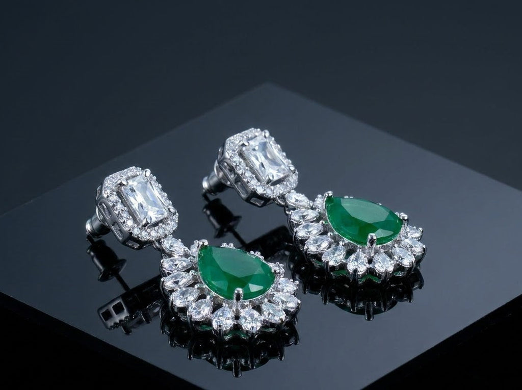 A pair of silver-toned drop earrings with oval green emerald-cut center stones and surrounding clusters of clear stones, displayed on a glossy black surface.