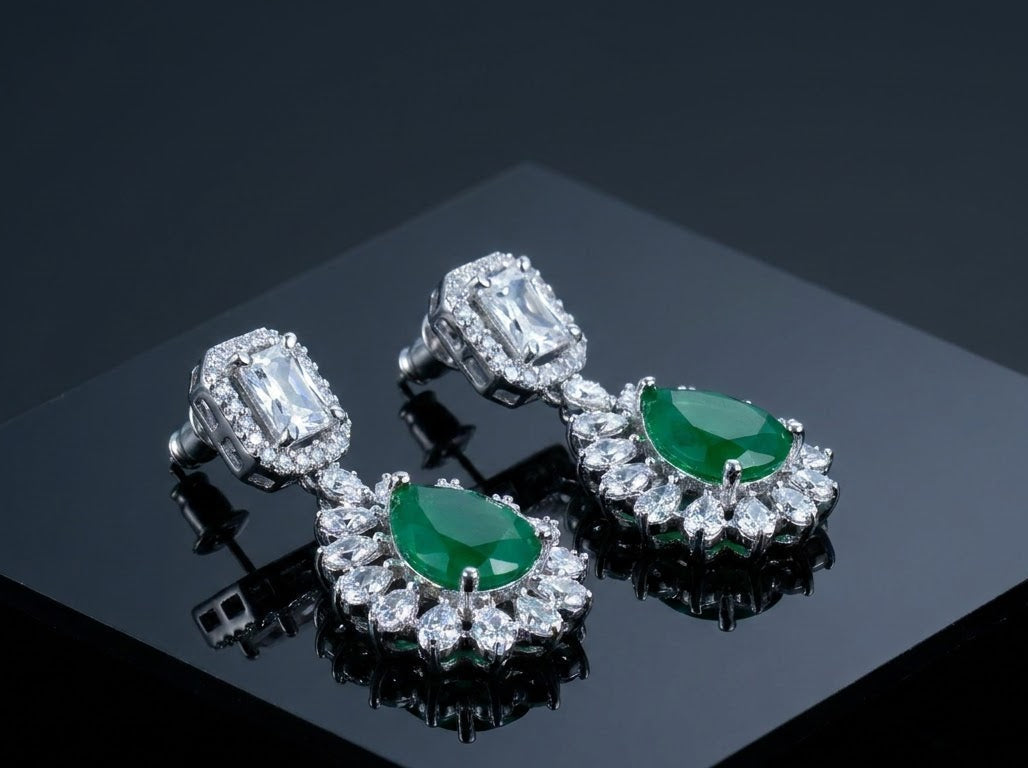 A pair of silver-toned drop earrings with oval green emerald-cut center stones and surrounding clusters of clear stones, displayed on a glossy black surface.