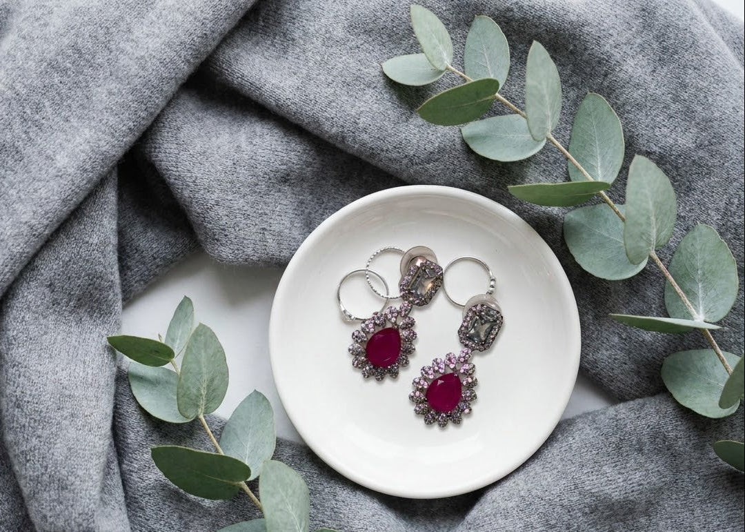 A pair of silver-toned statement earrings with deep red teardrop gemstones and textured silver halos, displayed on a small white dish surrounded by gray draped fabric and eucalyptus leaves.