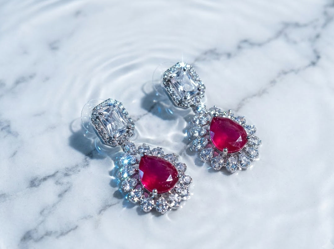 A pair of silver-toned drop earrings with large oval red gemstones surrounded by clear stones, resting on a white marble surface with water ripples.
