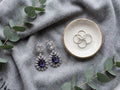 A pair of silver drop earrings with clear rectangular stones and deep purple teardrop-shaped center stones, displayed on a gray knit blanket next to a small white dish with gold trim holding three rings, with eucalyptus leaves around them.