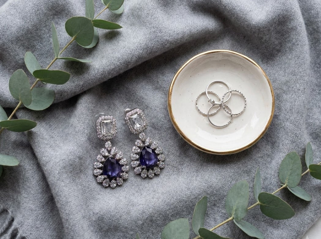 A pair of silver drop earrings with clear rectangular stones and deep purple teardrop-shaped center stones, displayed on a gray knit blanket next to a small white dish with gold trim holding three rings, with eucalyptus leaves around them.