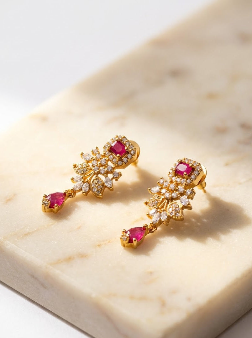 A pair of gold drop earrings with pink gemstones and clear crystal-like accents, displayed on a cream-colored marble slab against a white background.