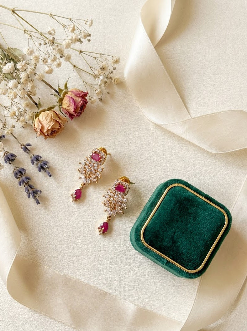 A pair of gold-toned statement earrings with pink gemstones and clear crystal-like details, arranged beside a small dark green velvet jewelry box with a gold border on a cream background with dried flowers and a cream ribbon.