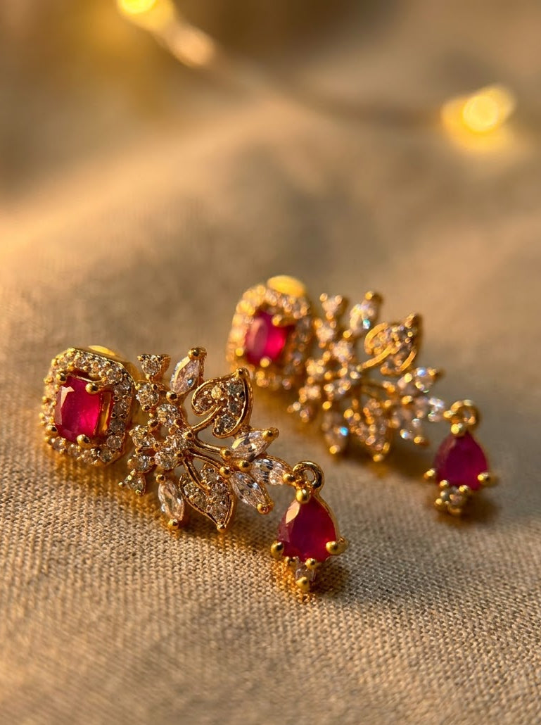 A pair of gold-toned floral chandelier earrings with square and teardrop-cut pink gemstones and clear crystal accents, displayed on a textured beige fabric background with warm bokeh lights.