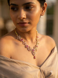 A woman wearing a gold-toned traditional necklace with pink gemstones and clear stones, styled with a beige draped garment.