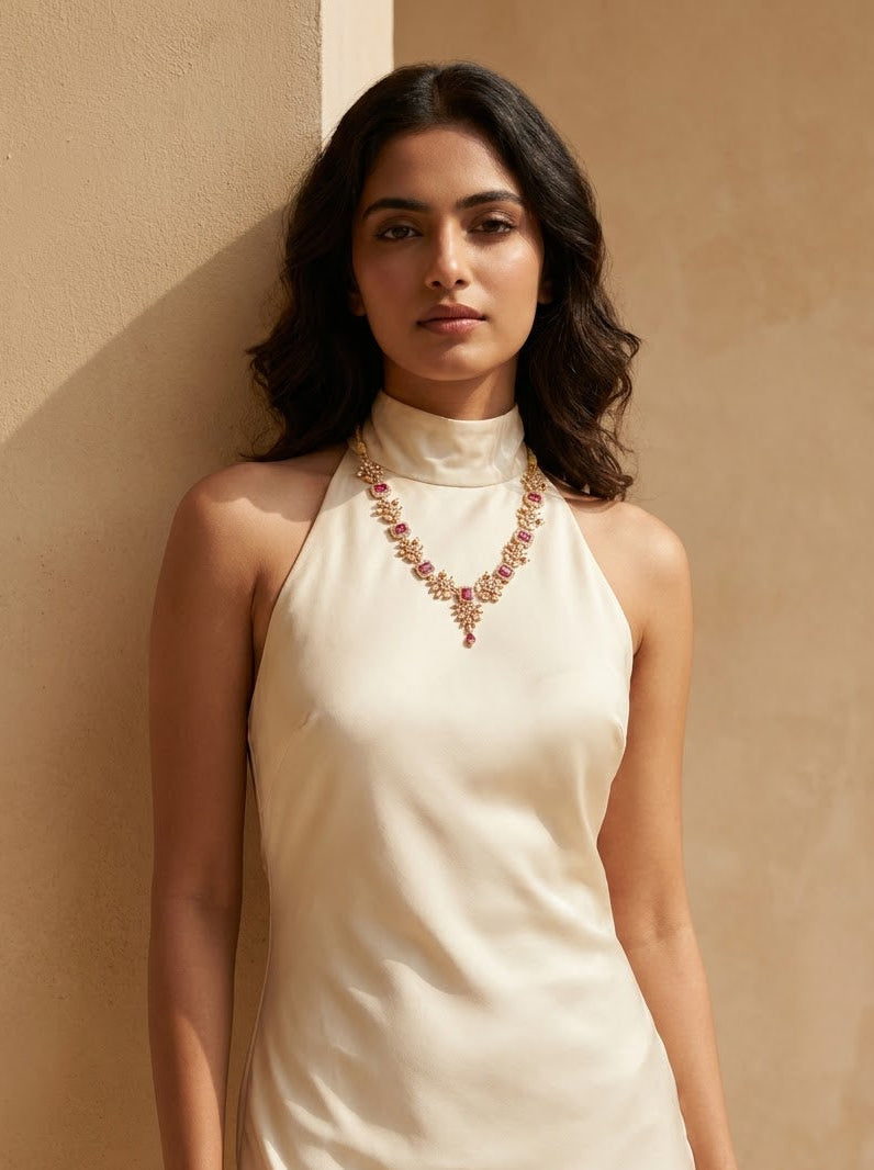 A woman wearing a cream-colored high-neck sleeveless dress with a decorative Ruby and gold embroidered necklace, standing against a textured beige wall.