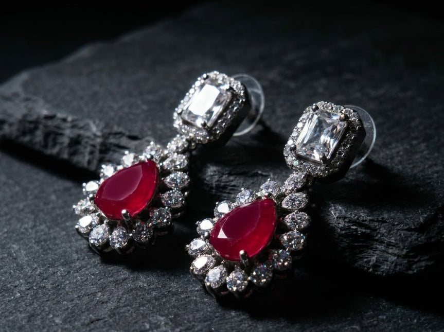 A pair of silver-toned dangle earrings featuring marquise-cut red gemstones surrounded by clear stones, displayed on a dark textured stone surface.
