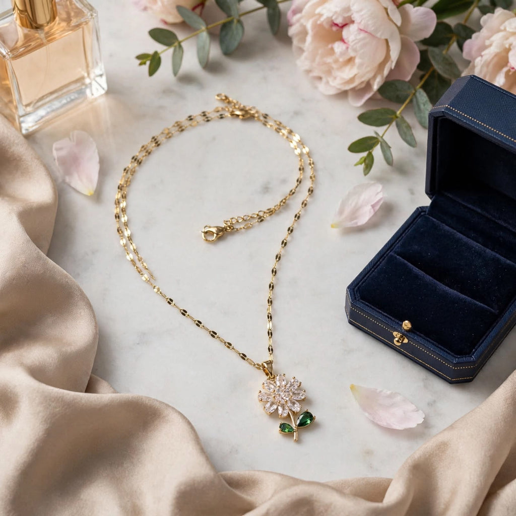 A gold pendant necklace with a flower and green leaf design lies on a white marble surface, styled with a navy jewelry box, satin fabric, flowers, and a perfume bottle.