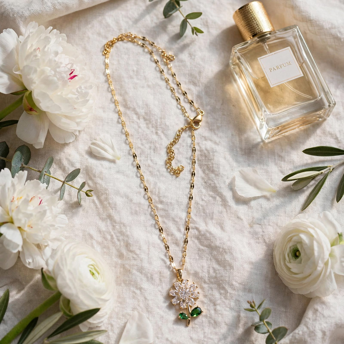 A gold chain necklace with a floral pendant featuring clear crystals and green gemstones, styled on a textured white fabric alongside white flowers, green leaves, and a perfume bottle labeled PARFUM.