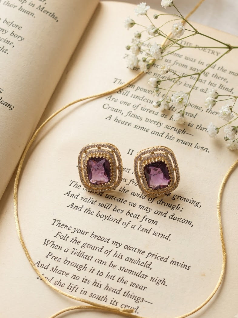 A pair of gold-toned stud earrings with rectangular purple gemstones, styled on an open vintage book with delicate white baby's breath flowers.