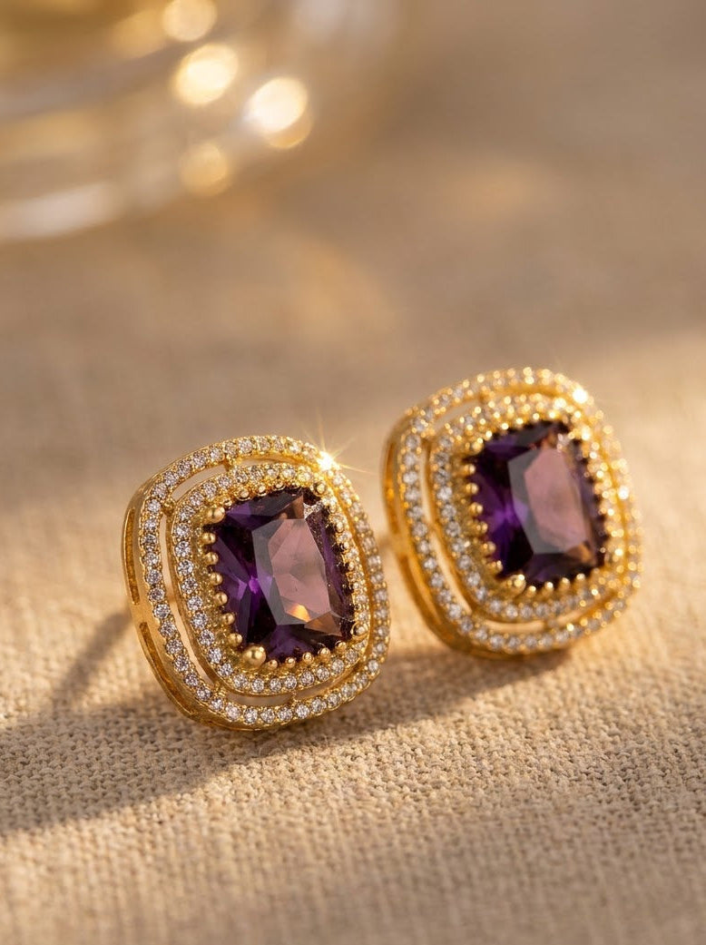 A pair of gold-toned stud earrings with cushion-cut purple gemstones and a halo of small clear stones, displayed on a textured beige fabric surface with warm golden bokeh in the background.