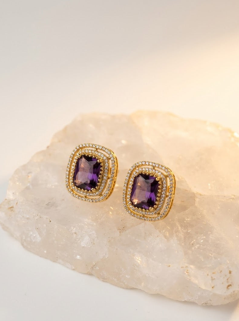 A pair of gold square stud earrings with rectangular purple amethyst centers and small clear stones around the edges, displayed on a pale crystal slab against a soft white background.