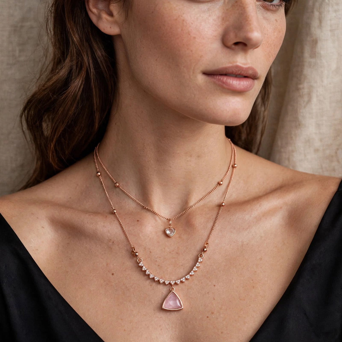 A close-up of a woman wearing a layered rose gold necklace set. The set includes a delicate chain with a small heart-shaped clear stone and a longer chain featuring a pink triangular pendant and a row of small pearls.