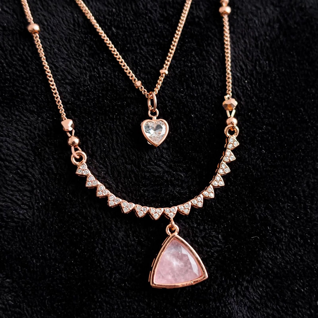 Rose gold layered necklace set on a black textured background, featuring a short chain with a clear heart-shaped crystal pendant and a longer chain with a triangle pendant set with small clear crystals.