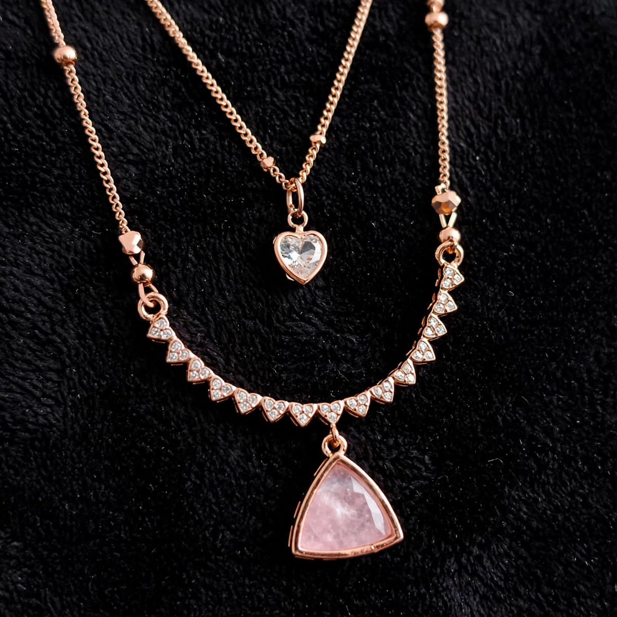 Rose gold layered necklace set on a black textured background, featuring a short chain with a clear heart-shaped crystal pendant and a longer chain with a triangle pendant set with small clear crystals.