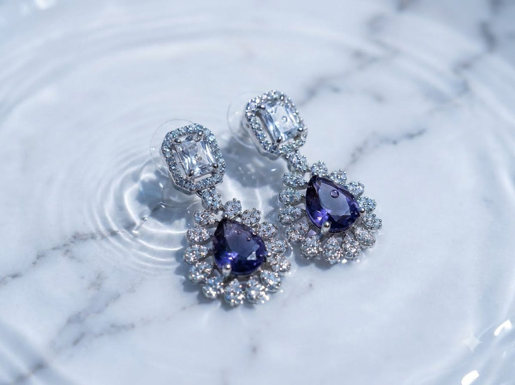 A pair of silver-toned dangle earrings with large pear-cut purple gemstones surrounded by clear crystals, resting on a white marble surface with water ripples.