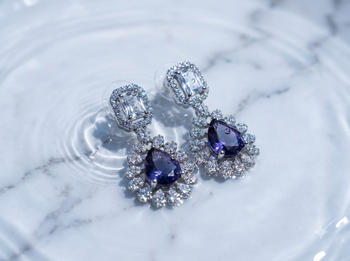 A pair of silver-toned dangle earrings with large pear-cut purple gemstones surrounded by clear crystals, resting on a white marble surface with water ripples.