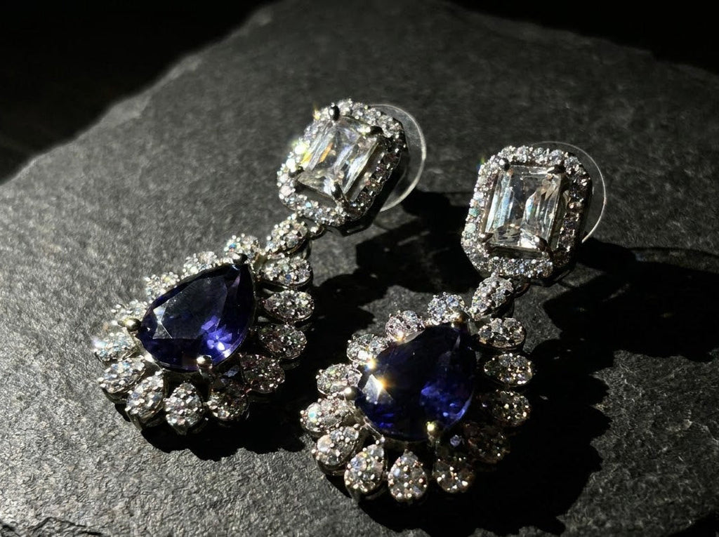 A pair of silver-toned drop earrings featuring a large teardrop-shaped blue sapphire and a rectangular clear gemstone, surrounded by small clear stones in a floral setting, displayed on a dark textured stone surface.