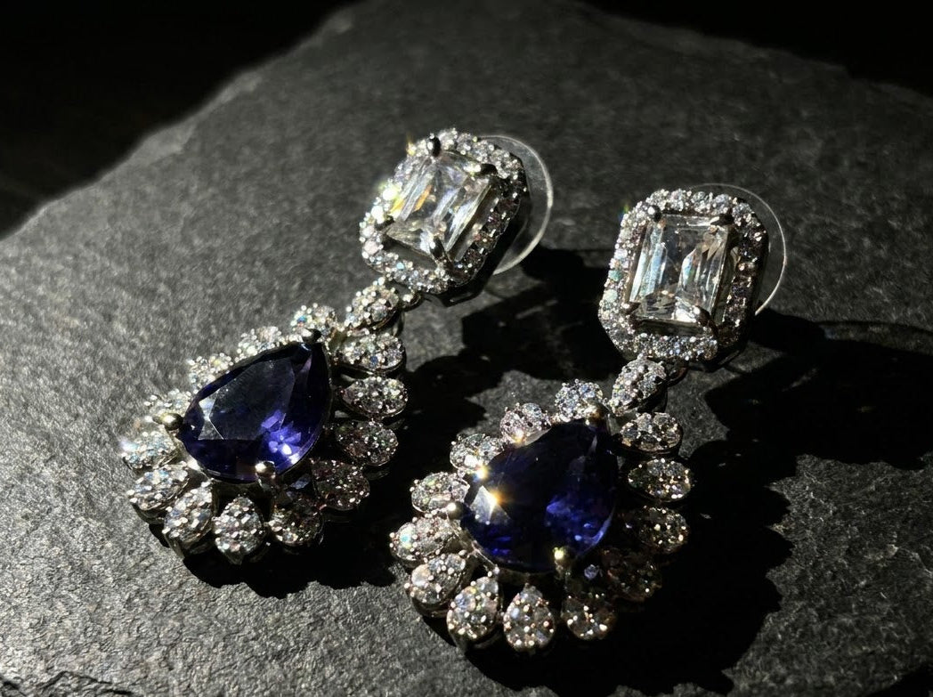 A pair of silver-toned drop earrings featuring a large teardrop-shaped blue sapphire and a rectangular clear gemstone, surrounded by small clear stones in a floral setting, displayed on a dark textured stone surface.
