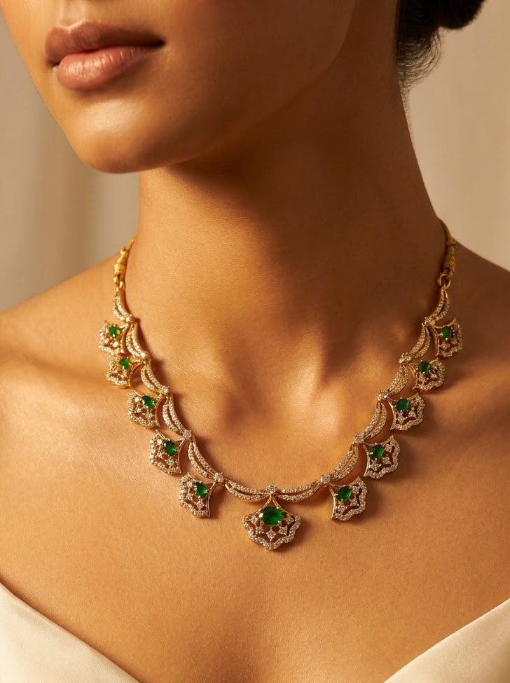 A close-up of a model wearing a gold statement necklace adorned with green emeralds and small clear stones, paired with a cream satin top against a warm beige background.