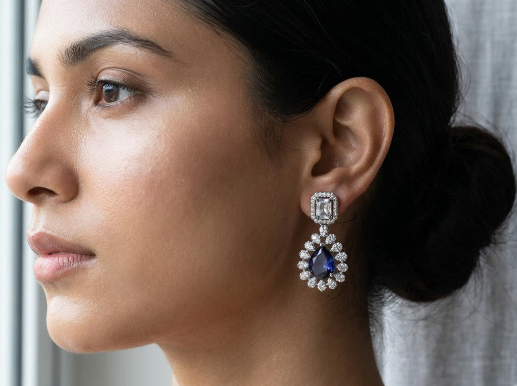 A close-up side profile of a woman wearing a silver-toned dangle earring with a rectangular clear gemstone and a teardrop-shaped blue sapphire surrounded by small clear stones, set against a soft gray curtain background.