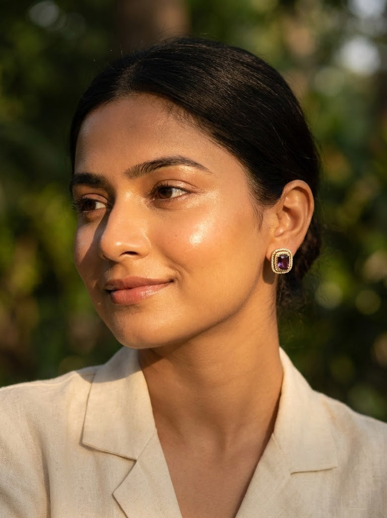 A woman with dark hair pulled back wears a cream-colored blazer and a small gold-toned earring with a purple gemstone, set against a soft-focus green outdoor background.