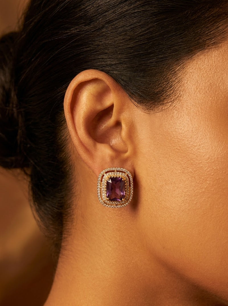 A close-up side profile of a woman wearing a single rectangular stud earring with a purple gemstone surrounded by a halo of small clear stones in a gold-tone setting.