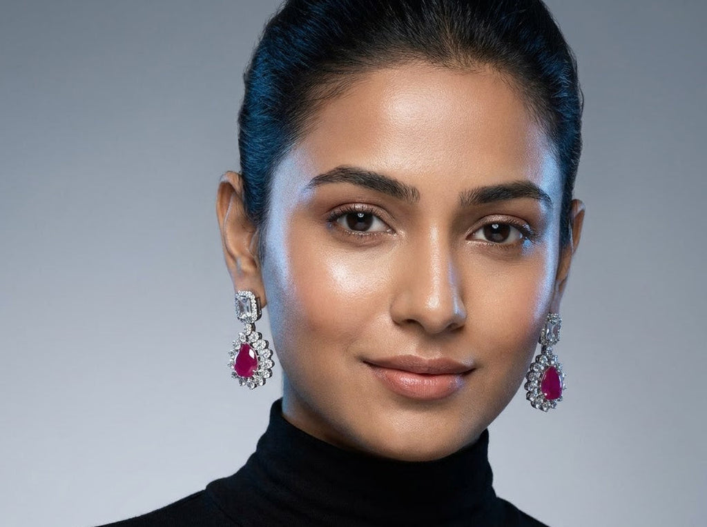 A close-up portrait of a woman wearing a black turtleneck sweater and matching drop earrings with bright pink teardrop-shaped stones and silver-toned settings, against a soft gray studio background.