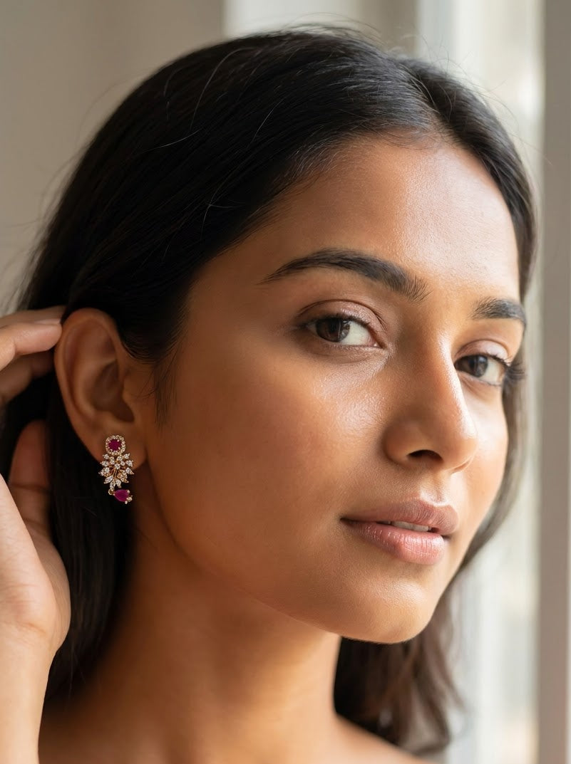 A close-up portrait of a woman wearing a small, ornate gold-toned earring with a pink gemstone, set against a soft, bright window background.