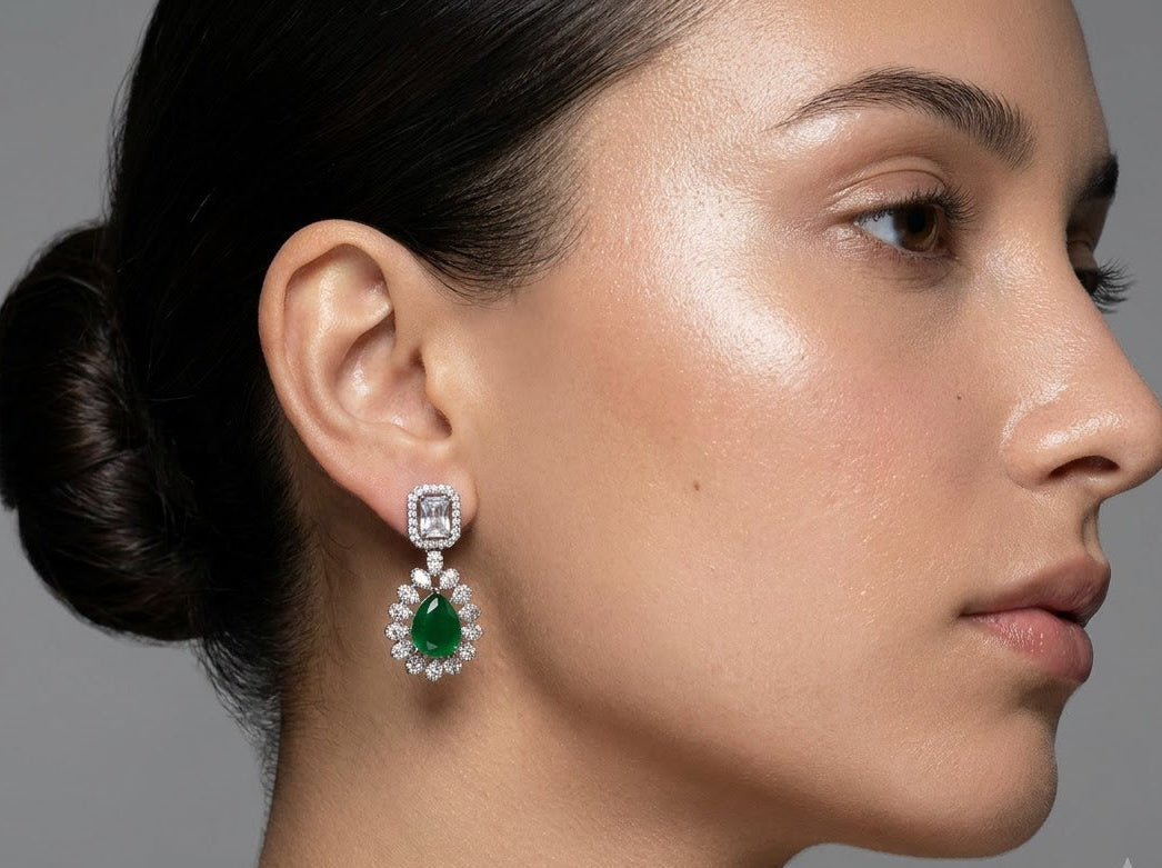 A close-up side profile of a woman wearing a dangling earring. The earring features a square clear gemstone in a silver-toned setting with a teardrop-shaped green gemstone surrounded by small clear stones below it, set against a neutral gray background.