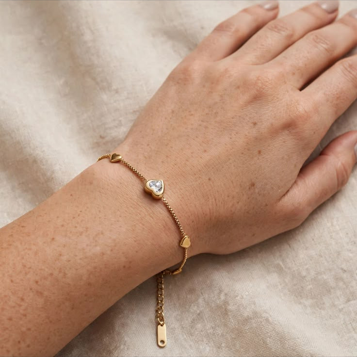 A delicate gold chain bracelet with a small heart-shaped crystal charm and a dangling tag, shown worn on a person's wrist against a soft beige fabric background.